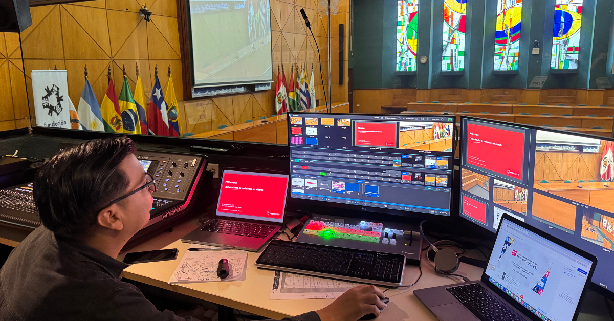 Photo featuring a person monitoring multiple computer screens and a sound controller during a conference talk. From the control room, there is a view to a wooden paneled lecture hall, with 14 flags of Latin American countries on stage and a stained glass window to the right side of the hall.